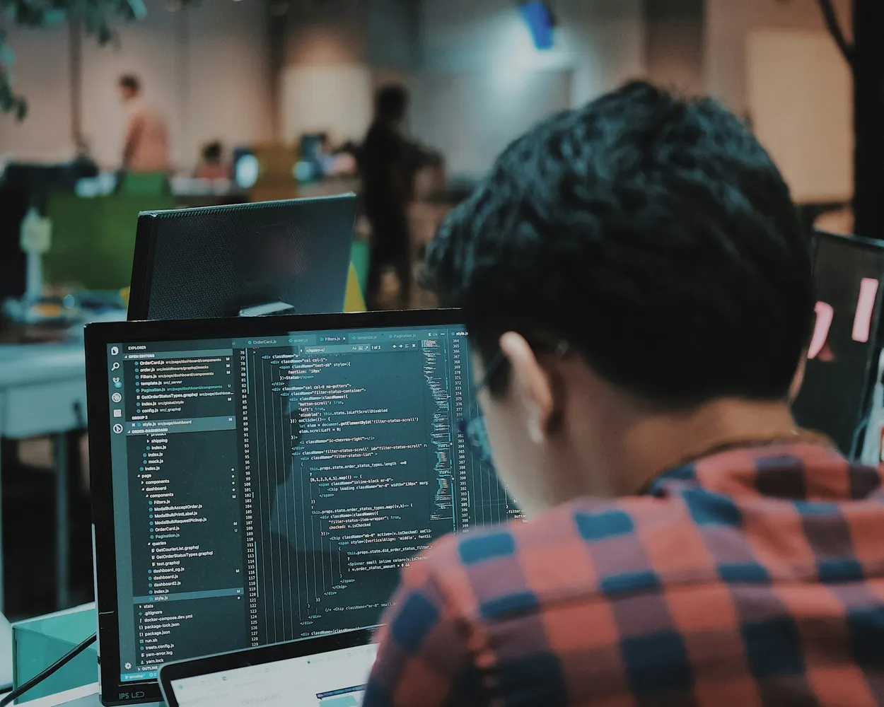 Programador visto de costas num escritório, com o código de uma aplicação aberto num monitor e um portátil à frente